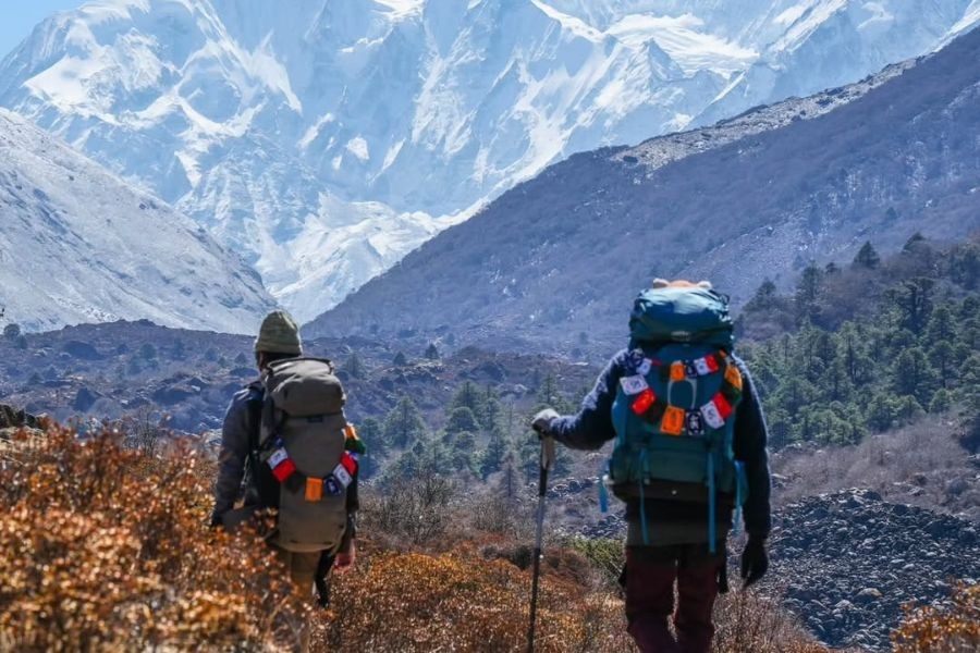 Two trekkers with backpacks walking on mountain trail with snowy peaks in background